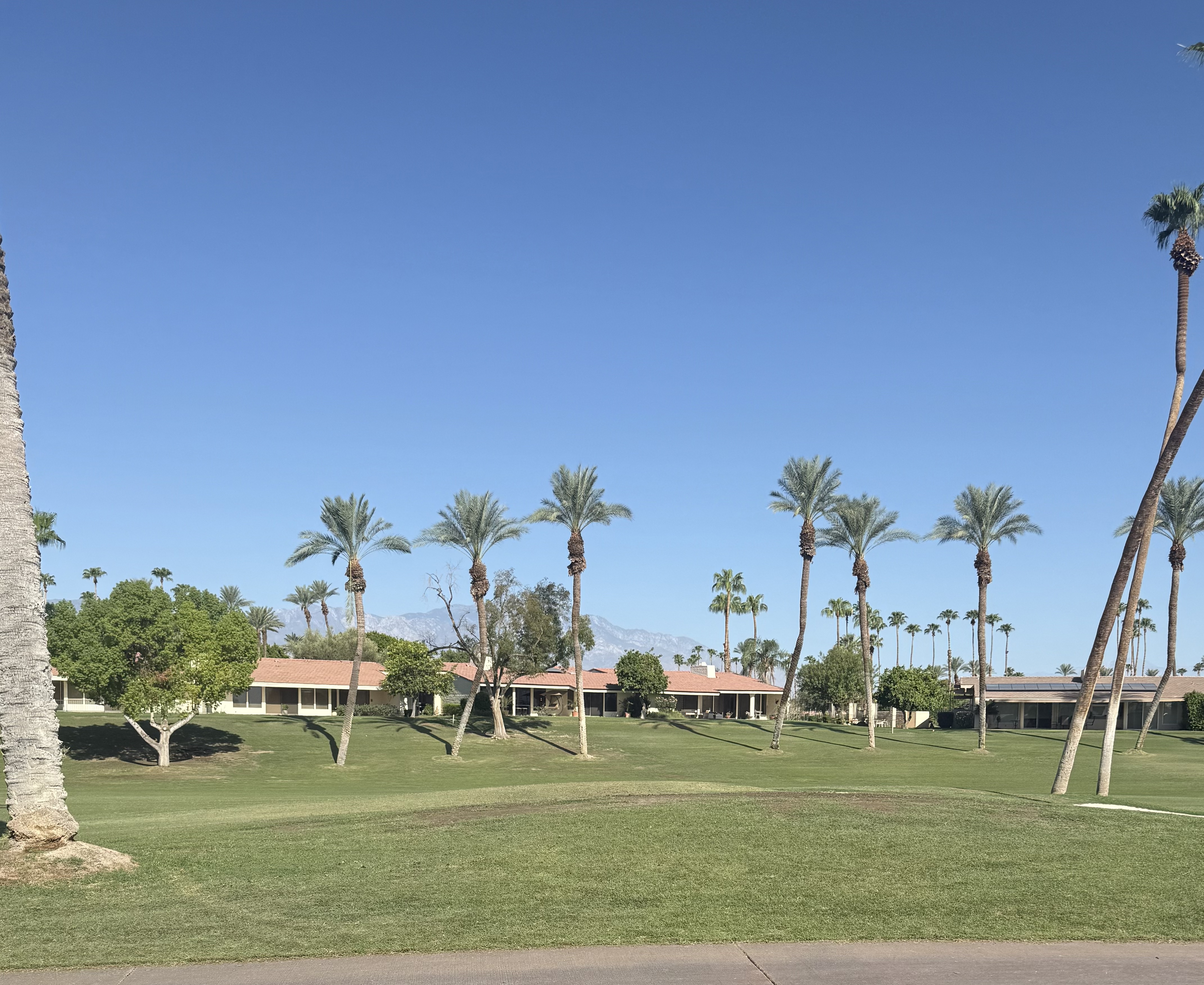 An aerial photographic view of a Coachella Valley master-planned community, showing a mosaic of tan-roofed homes, curved cul-de-sacs, and meandering green fairways of a golf course set against the surrounding arid landscape. Blue community pools, tidy walking paths, and pockets of palm-lined parks add color and structure. The image is captured in bright midday sunlight, rendering clear details and minimal shadows for a factual, map-like clarity. Shot from directly overhead with a wide frame, the composition emphasizes patterns and density, creating a professional, data-driven visual suited to articles about development trends and community growth.